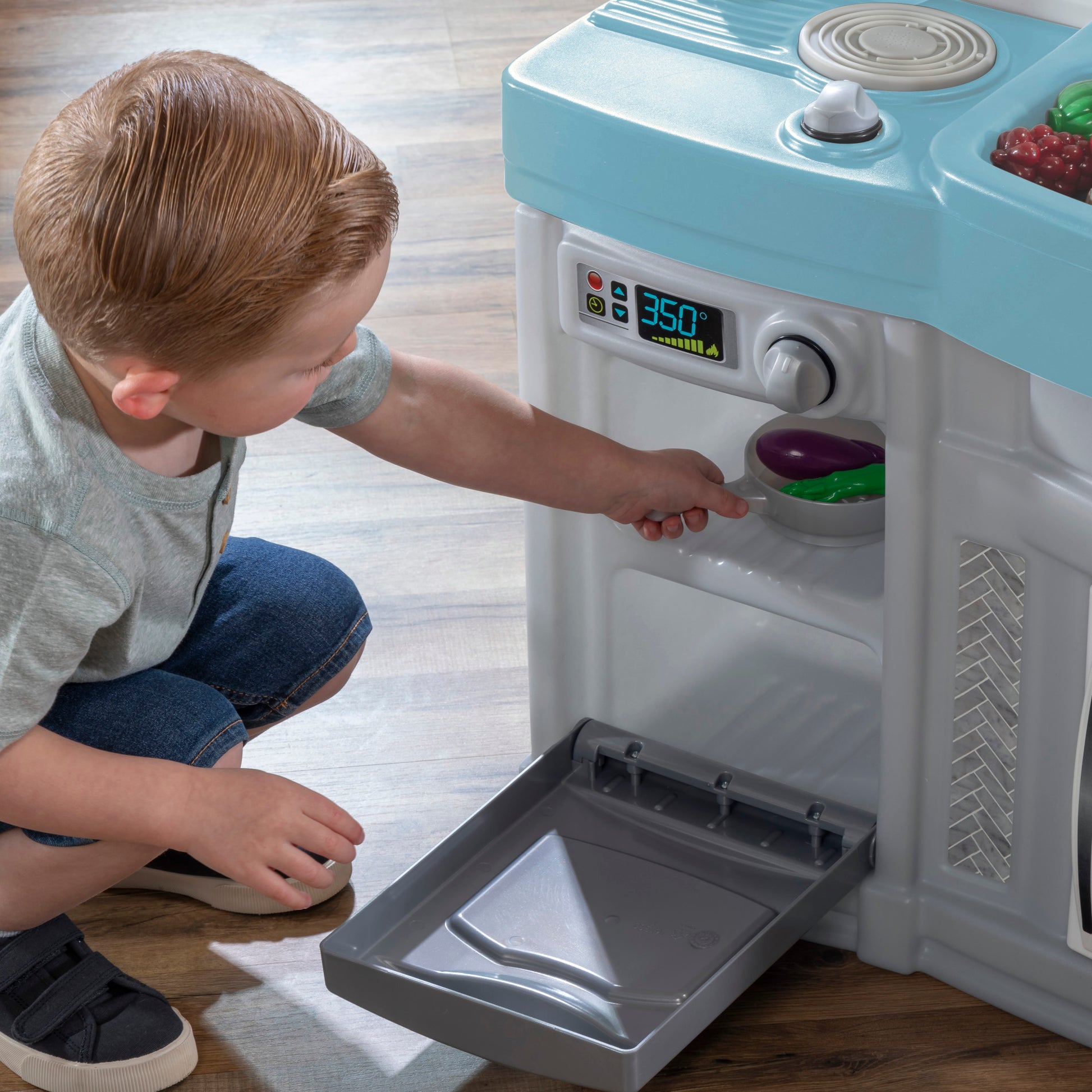 Choice Cuisine Kitchen — boy crouching at gray oven door, placing toy eggplant inside; digital display reads 350 degrees; light blue countertop above