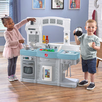 Choice Cuisine Kitchen — girl in pink dress reaching to stovetop and boy in green shirt holding toy coffeemaker; both smiling; blue and gray toy kitchen in indoor playroom setting