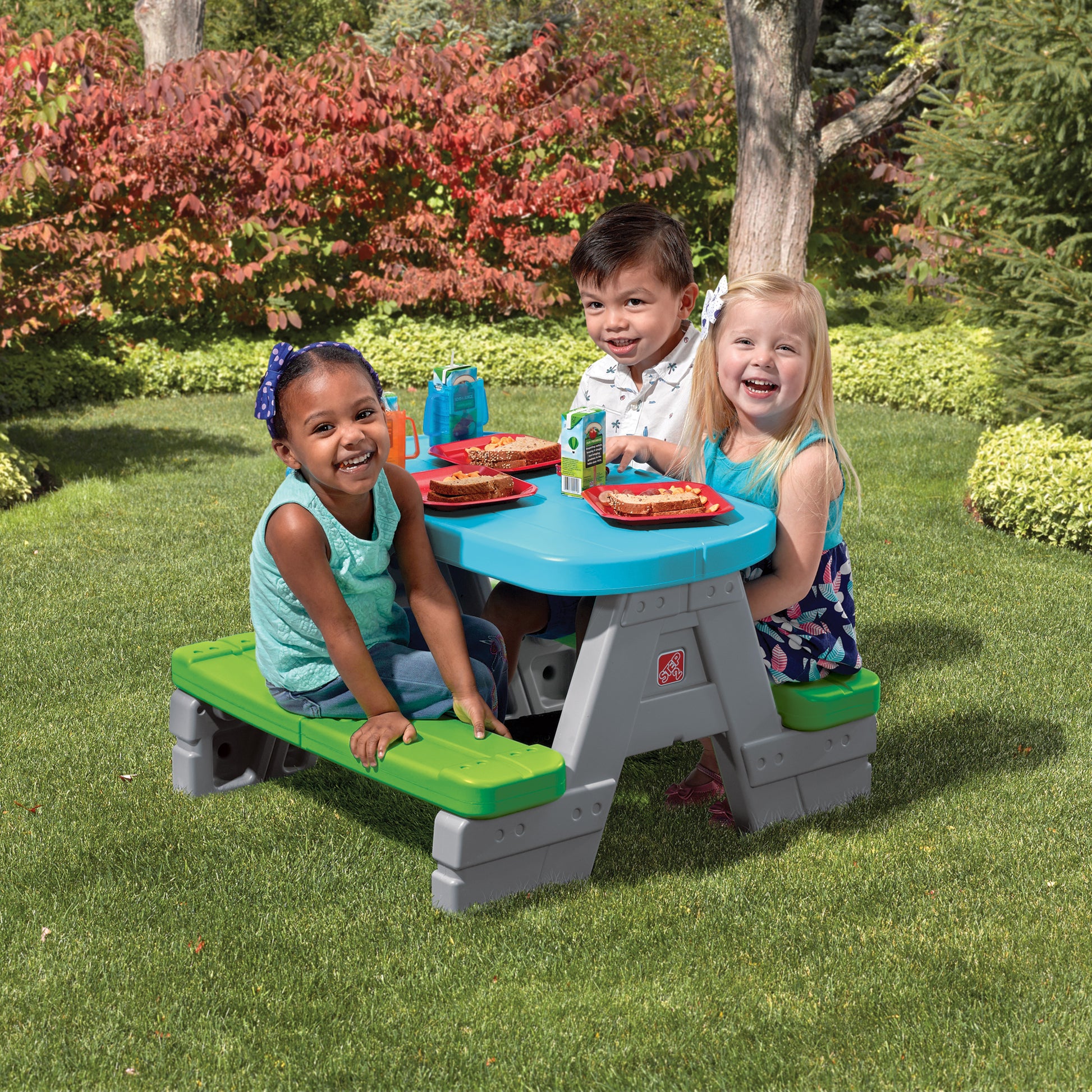 Fun & Friends Picnic Table™ being enjoyed by three toddler children eating at blue picnic table on grass with red plates, food, and juice boxes; colorful fall foliage background