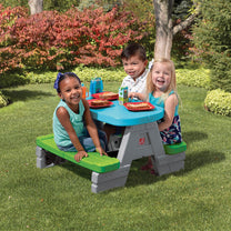Fun & Friends Picnic Table™ being enjoyed by three toddler children eating at blue picnic table on grass with red plates, food, and juice boxes; colorful fall foliage background