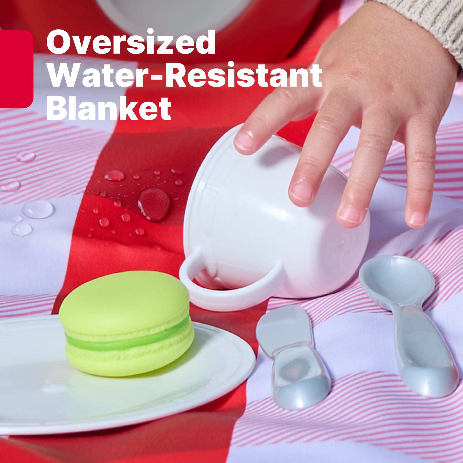 Oversized Water-Resistant Blanket — close-up of child's hand pouring toy tea; green macaron and toy spoons on red-and-white striped blanket with water beading off
