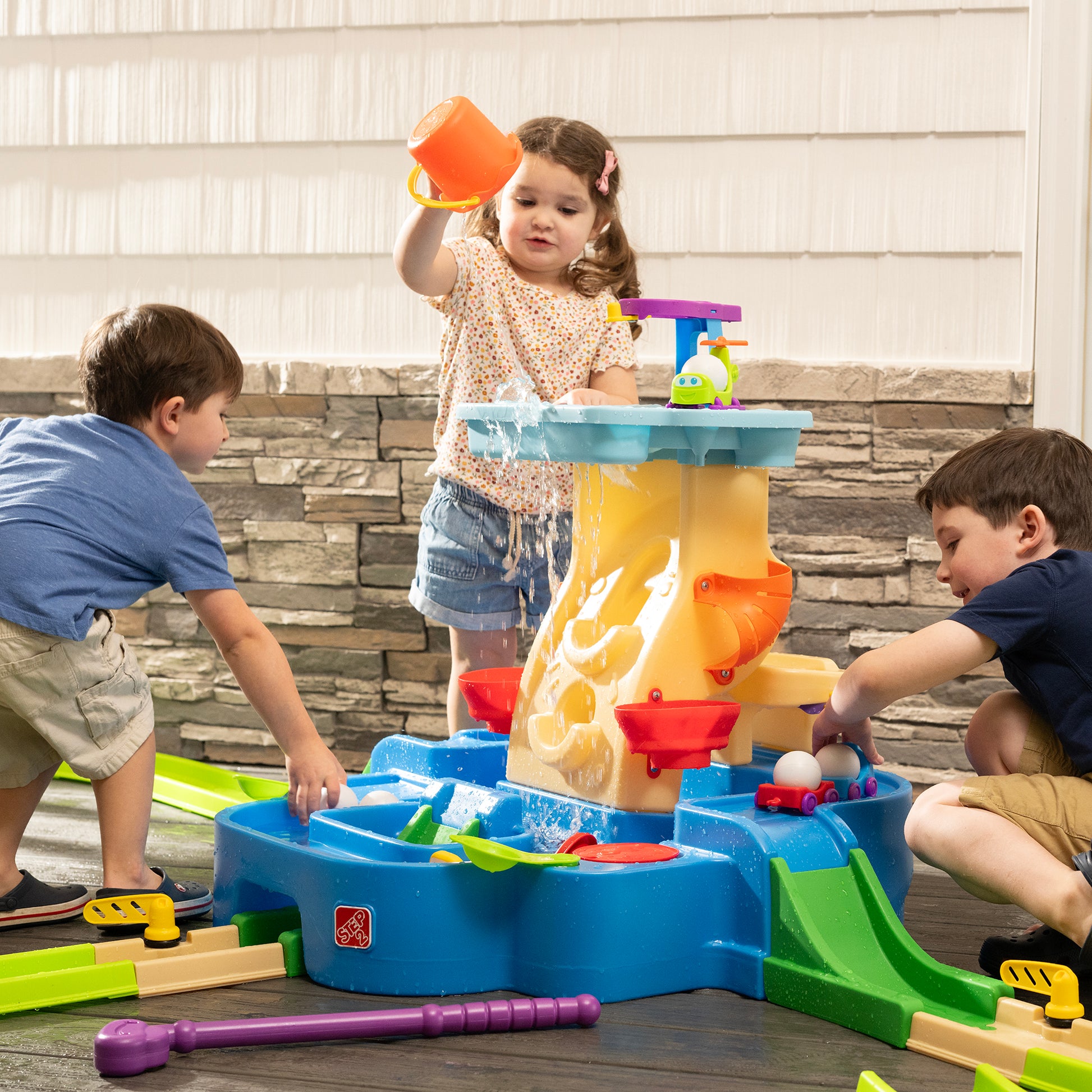 Putt-Splash Adventure Center — three children playing indoors; girl pouring orange cup over water tower causing splash; boys on each side reaching into water basin; stone wall backdrop