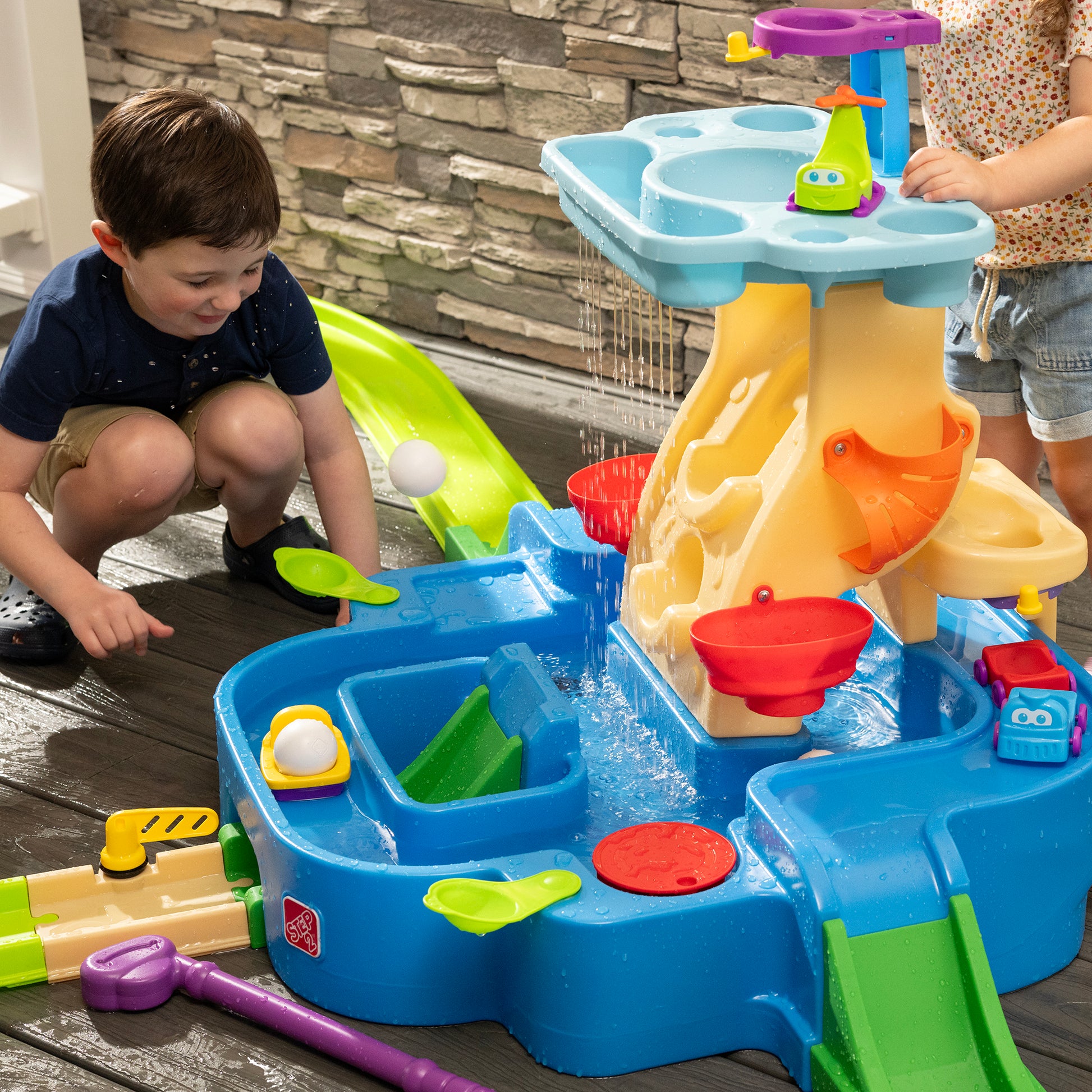 Putt-Splash Adventure Center — boy crouching at water basin watching toy vehicles float and water cascade down tan tower; second child visible; blue water basin with colorful accessories