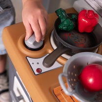 Close-up overhead view of child's hand turning the Garden Fresh Kitchen™ burner knob; dark gray frying pan with play broccoli and red pepper on burner; gray colander with red tomato beside