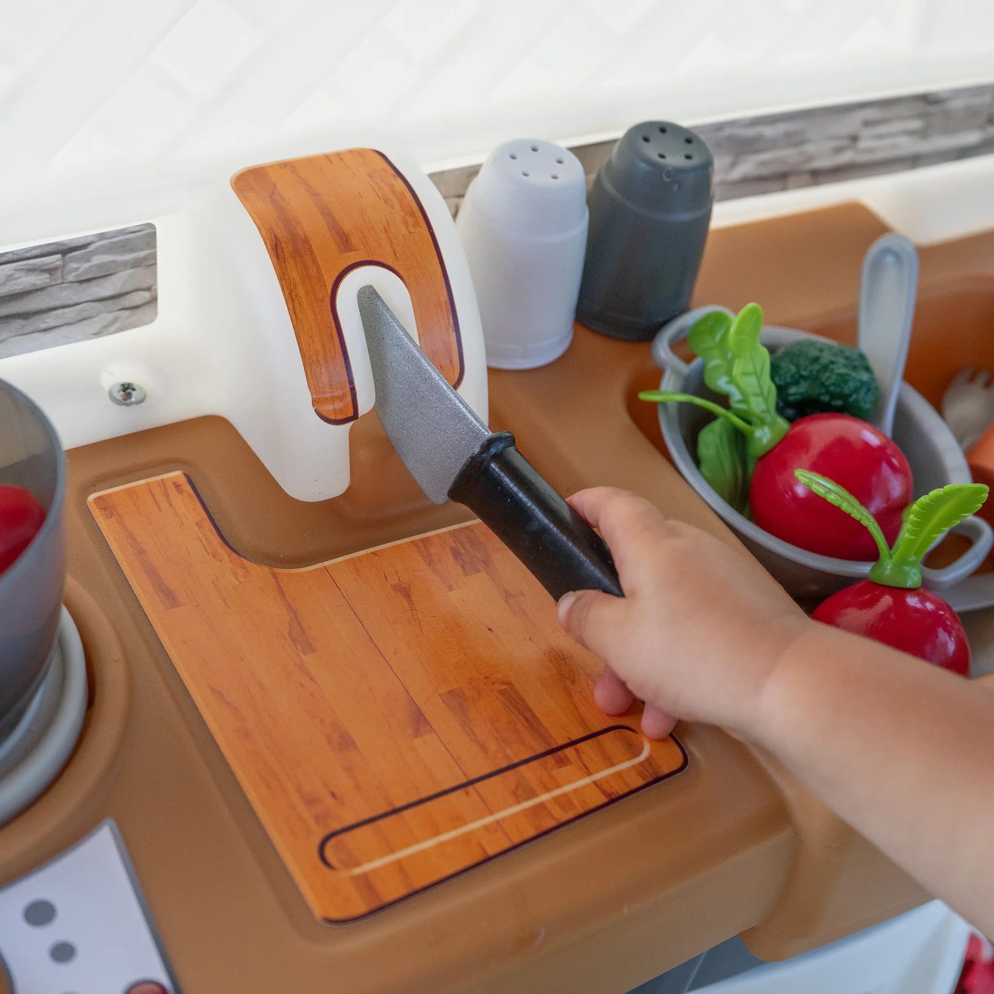 Close-up of child's hand using a play knife on a wooden cutting board at the Garden Fresh Kitchen™; knife holder, salt and pepper shakers, gray colander with red tomato and broccoli visible on the butcher block countertop