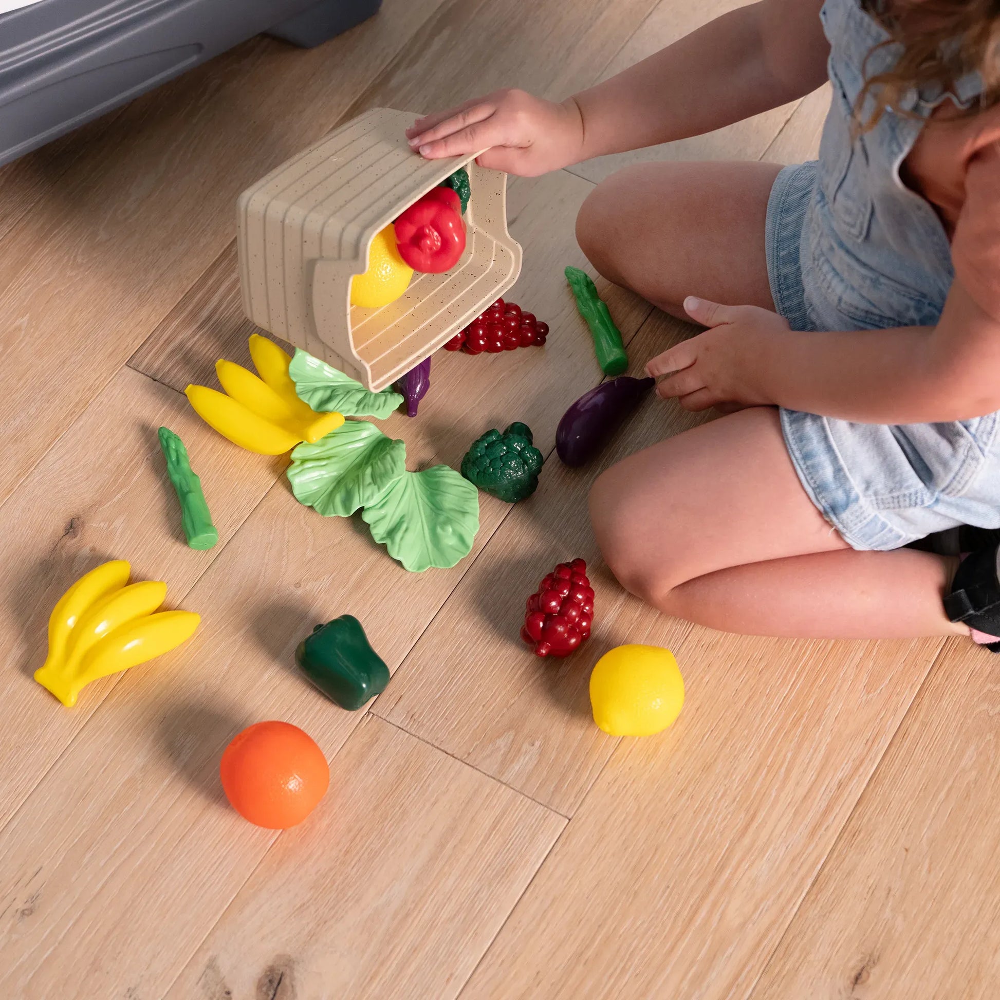 Garden Fresh Kitchen™ being enjoyed by toddler girl sitting on the floor, emptying a produce crate; 13+ play food pieces scattered including bananas, peppers, grapes, eggplant, asparagus, lettuce, orange, and broccoli