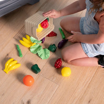 Garden Fresh Kitchen™ being enjoyed by toddler girl sitting on the floor, emptying a produce crate; 13+ play food pieces scattered including bananas, peppers, grapes, eggplant, asparagus, lettuce, orange, and broccoli