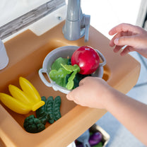 Close-up overhead view of child's hands placing a red apple in the gray colander in the Garden Fresh Kitchen™ sink basin; bananas, cucumber, and broccoli also visible in sink area