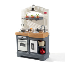 Step2 Garden Fresh Kitchen™ — dark gray base with oven and refrigerator door panels, golden butcher block countertop, white arched backsplash with diamond tile texture, gray shelf with three herb pots under black pendant lamp; accessories include red watering can, cream basket, red pitcher; silver gooseneck faucet and sink; 3/4 front-left view on white background