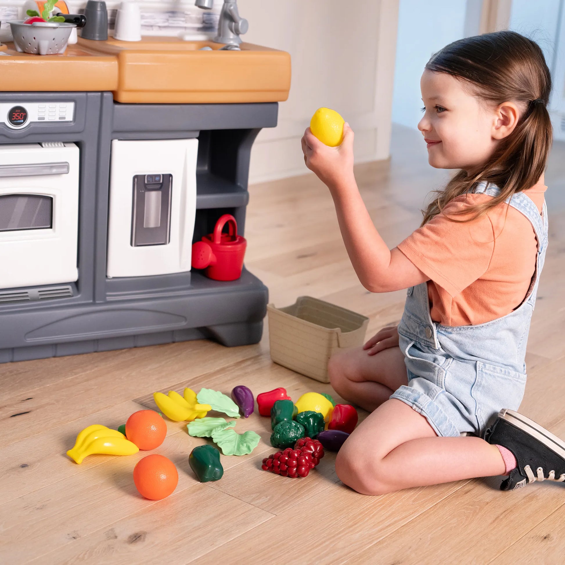 Garden Fresh Kitchen™ being enjoyed by girl in denim overalls and orange shirt kneeling on wood floor, holding a yellow lemon with play food spread in front; kitchen base visible behind