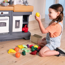 Garden Fresh Kitchen™ being enjoyed by girl in denim overalls and orange shirt kneeling on wood floor, holding a yellow lemon with play food spread in front; kitchen base visible behind