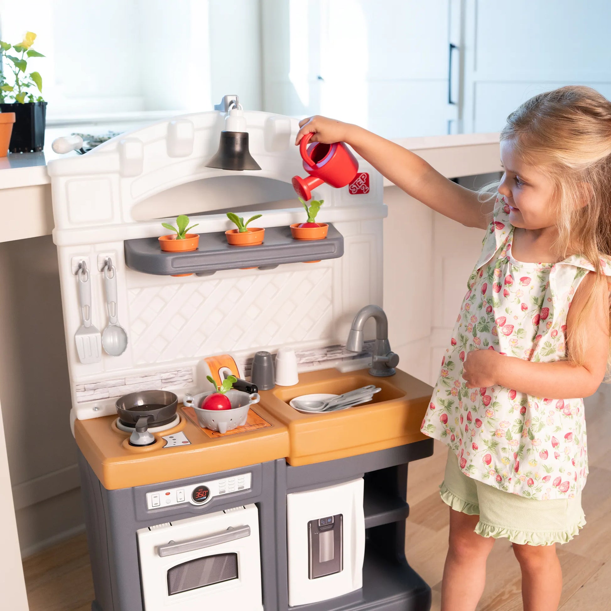 Garden Fresh Kitchen™ being enjoyed by blonde girl in strawberry-print dress watering herb pots with red watering can from above; full front kitchen view with real kitchen window background