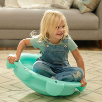 Toddler girl in overalls sitting cross-legged in Topsy Turtle teal rocking disk, holding hand grips, in living room