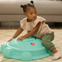 Toddler girl in beige blouse kneeling on top of Topsy Turtle teal rocking disk, using it as a stepping stone indoors