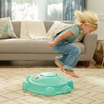 Toddler girl in overalls jumping off Topsy Turtle teal rocking disk in living room — hair in motion