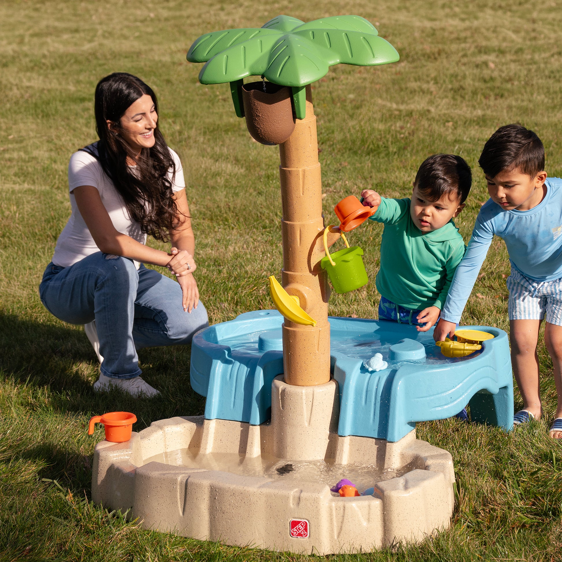 Tropical Splash Water Park™ being enjoyed by mom kneeling on grass watching two young boys play with water and accessories at the outdoor water table