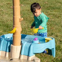 Tropical Splash Water Park™ being enjoyed by toddler boy in green hoodie pouring water from yellow sieve into the upper rock basin