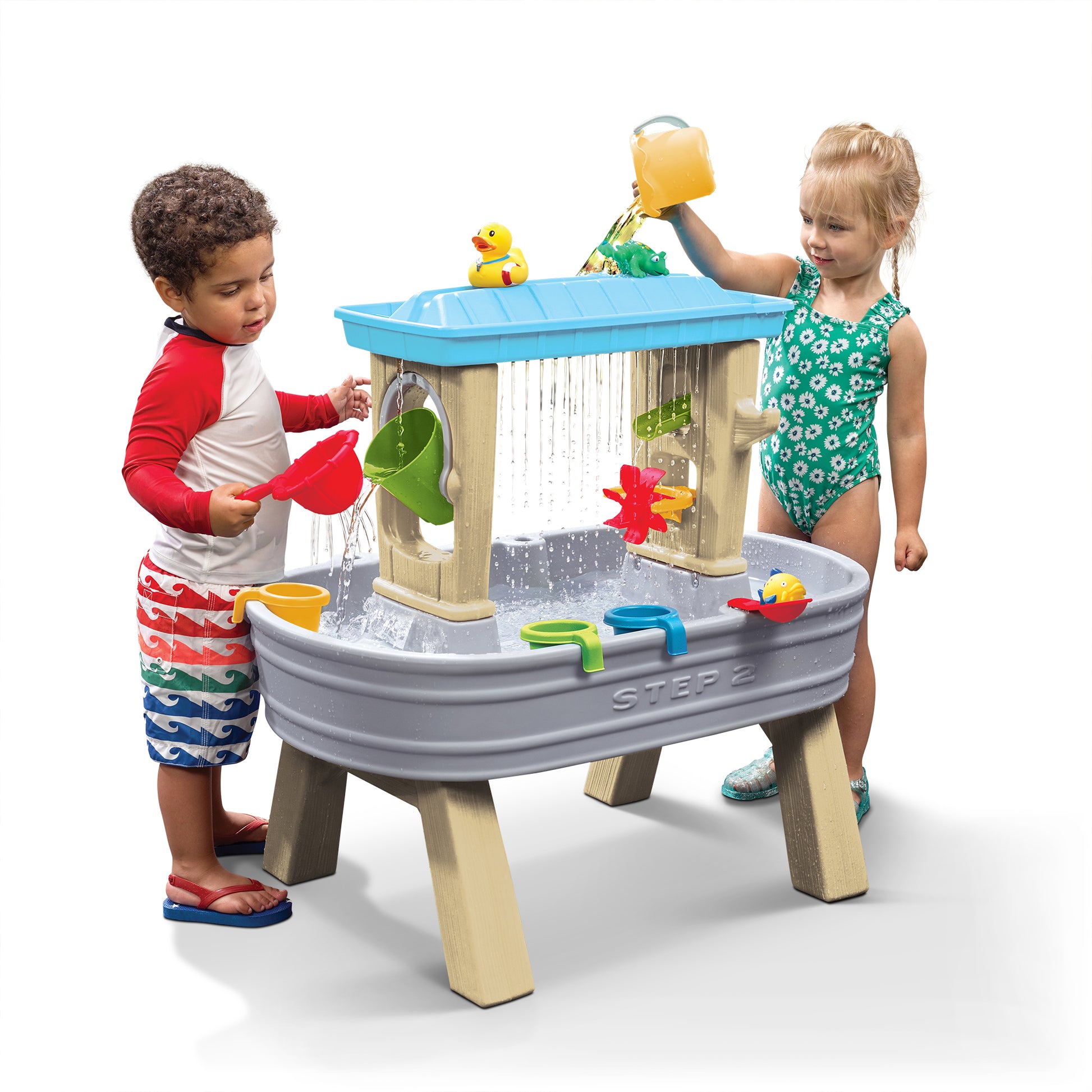 Step2 Rain Showers Splash Station Water Table™ — two toddlers in swimsuits playing at table with gray/stone-textured base, tan pillars, and blue upper rain tray; accessories include rubber duck, pinwheel, and cups; white background