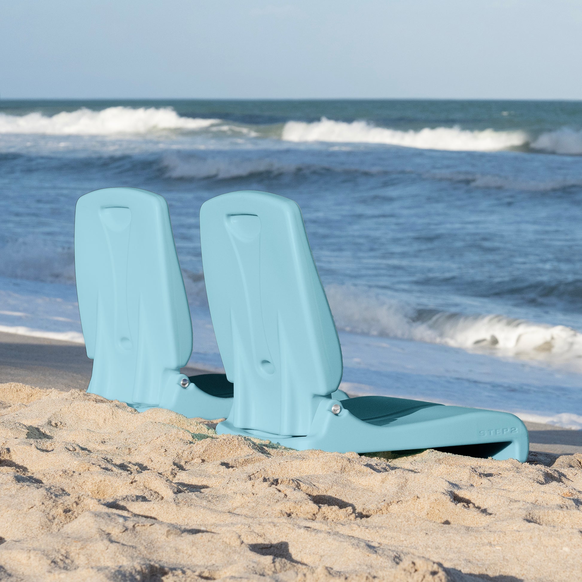 Two Flip Seat™ chairs in teal propped upright side by side in beach sand near the ocean shoreline