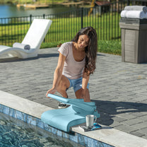 Woman kneeling at pool edge unfolding a teal Flip Seat™ with books and a mug on the nearby ledge