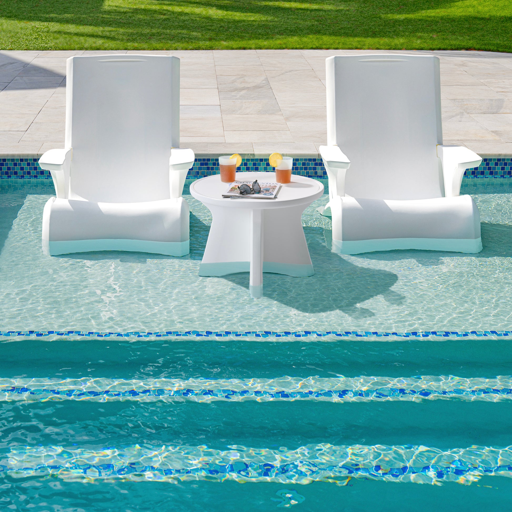 Vero In Pool Accent Table™ in shallow pool water between two white Vero Adirondack chairs — drinks and a magazine on tabletop; stone pool coping and lawn visible beyond