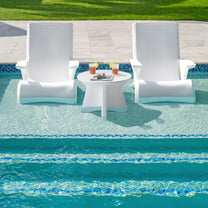 Vero In Pool Accent Table™ in shallow pool water between two white Vero Adirondack chairs — drinks and a magazine on tabletop; stone pool coping and lawn visible beyond