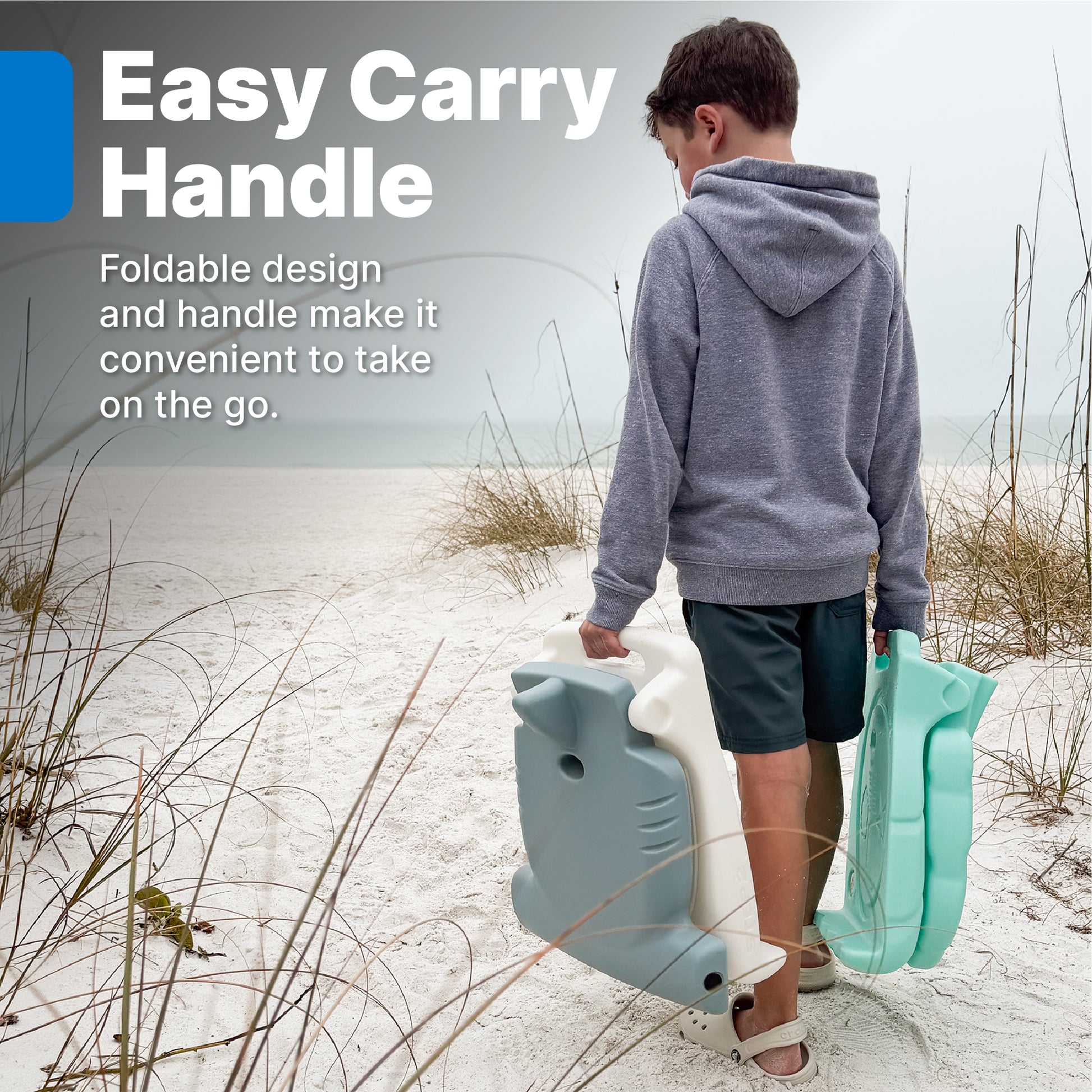 Kids Seashell Flip Seat — boy carrying folded teal flip seat and blue hammerhead seat on beach with dunes in background