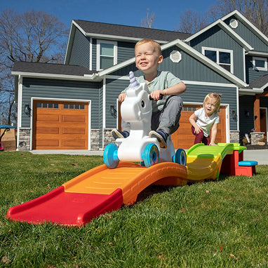 Children playing with a colorful toy slide and horse in front of a house.