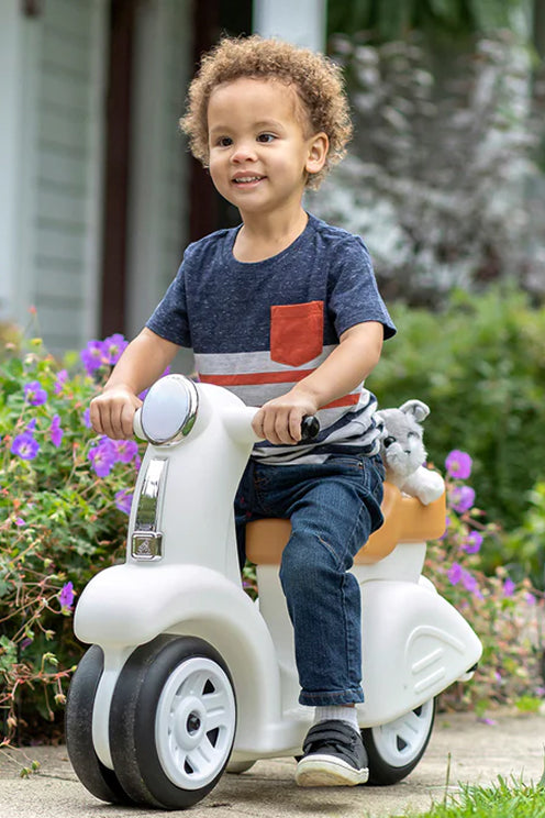 Child riding a toy scooter with a teddy bear on a garden path